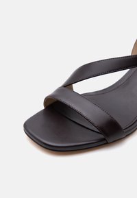 Μαύρο δερμάτινο σανδάλι με δύο φαρδιές λουράκια που διασταυρώνονται και τετράγωνη ανοιχτή μύτη σε λευκό φόντο.