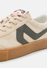 Beige velúr sneaker, sötétzöld zig-zag díszítéssel, texturált gumi talppal és bézs cipőfűzőkkel. Logó címke a nyelvén.