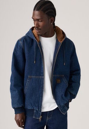 Uomo con capelli intrecciati che indossa una giacca di jeans blu con cappuccio sopra una camicia bianca, guarda verso il basso e di lato con una mano in tasca.