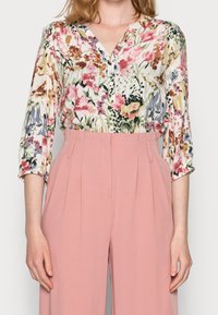 Kvinna som bär en blommig knäppt blus med 3/4-långa ärmar, inskjuten i högmidjade, veckade rosa vida byxor.