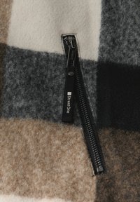 Fleecový oděv v béžové, šedé a hnědé kostkované vzoru; má černý zip s logem. Texturovaný povrch s jemným povrchem.