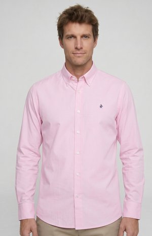 Hombre con una camisa de botones de color rosa claro con un pequeño logo oscuro bordado en el pecho, de pie frente a un fondo gris liso.