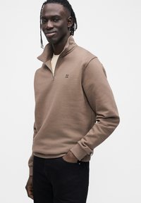 Brązowa bluza zapinana na zamek z wysokim kołnierzem, ściągaczami na rękawach i małym logo „II” na piersi, w połączeniu z czarnymi spodniami.