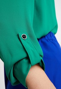 Grön blusärm med manschett som har en dekorativ svart ädelstensknapp och silverdetaljer; slät tyg med en lätt lyster.