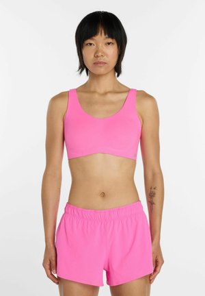 Kvinde med kort sort hår iført en knaldpink sports-bh og matchende shorts, stående mod en ensfarvet hvid baggrund.