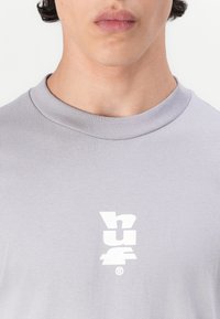 Lys grå T-shirt med rund hals, der har et hvidt grafisk logo foran. Glat tekstur og standard pasform.