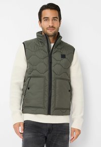 Steppweste in Olivgrün mit hohem Kragen, Frontreißverschluss und zwei großen Taschen. Getragen über einem cremefarbenen Strickpullover, der zu dunklen Jeans passt.