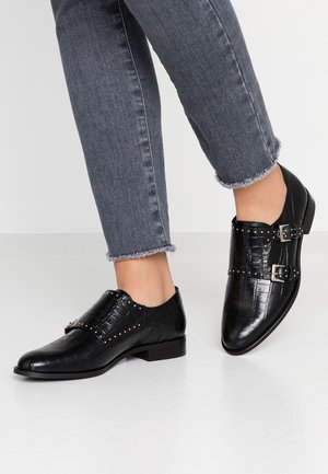 Zwarte structuurlederen loafers met zilveren gespen en studs, gedragen met grijze jeans met uitgefranjerde zoom op een lichtgrijze vloer.