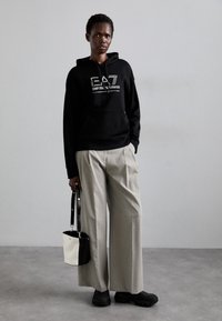 Svart hettegensere med logo, lysegrå plissert bukser, svart bøtteveske med hvite detaljer, og svarte chunky sneakers. Flat bakgrunn.