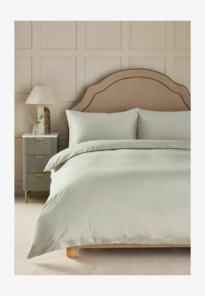 Bett mit beigem, geschwungenem Kopfteil, hellgrauer Bettwäsche und passenden Kissen, neben einem Nachttisch mit Lampe und kleiner Pflanze.