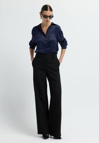 Chemise bleu marine en satin avec des manches retroussées, associée à un pantalon noir taille haute à jambes larges et des chaussures à talons noires. Lunettes de soleil portées.