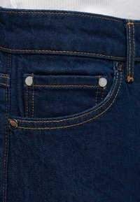 Τσέπη από σκούρο μπλε τζιν με χαλκομαντεμένα και πορτοκαλί ραφές σε λευκό υφασμάτινο φόντο.