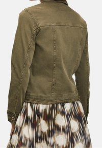 Chaqueta vaquera verde oliva con diseño ajustado, dobladillo desflecado, puños abotonados y costura en la espalda, combinada con una falda estampada.