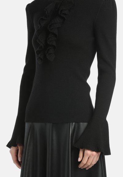 Pulover negru din tricot, cu un front volanat, textură cu nervuri și manșete evazate. Purtați deasupra unei fuste din piele pliată negre.