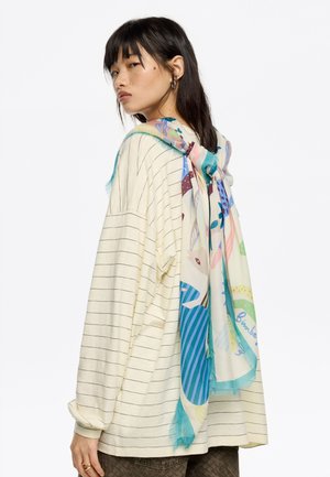 Bufanda de seda multicolor con un patrón juguetón, que presenta azules, verdes y rosas, drapeada sobre una camisa oversized con rayas color crema.