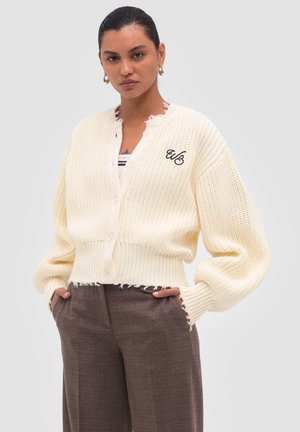 Vrouw draagt een crèmekleurige gebreide cardigan met zwarte "WB" borduursel, bruine broek, staat met handen in de zakken tegen een effen achtergrond.