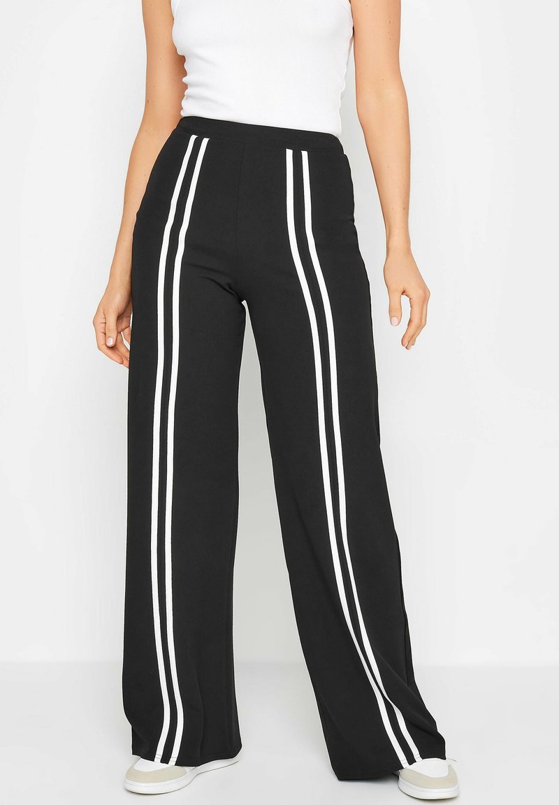 Zwart wijde broek met witte zijstroken, met een hoge taille en zachte stof. Gecombineerd met een witte top en lichtgekleurde sneakers.