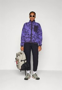 Violeta flīsa jaka ar ziedu rakstu, melna kabata, melnas bikses, pelēks mugursoma un pelēki zaļi apavi. Izskatu noslēdz saulesbrilles.