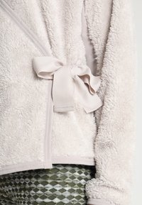 Jasnoszara puszysta kurtka z polaru związana szeroką tkaninową kokardą w talii, noszona na wzorzyste oliwkowe spodnie w romby.