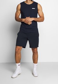 Top senza maniche blu scuro con un piccolo etichetta logo, abbinato a pantaloni corti blu scuro. Sneakers atletiche bianche e calzini bianchi alti con logo. Sfondo semplice.