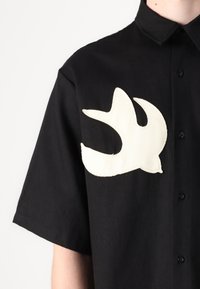 Camisa preta de manga curta com um destacado patch branco em forma de pássaro no lado esquerdo do peito. Feita de um tecido texturizado com fecho de botão.