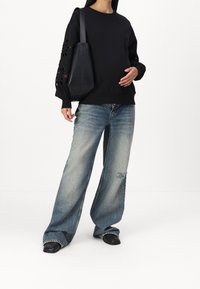 Schwarzer Pullover mit floralen Ausschnitten an den Ärmeln, kombiniert mit ausgefransten, weit geschnittenen blauen Jeans und schwarzen Slippern, dazu eine schwarze Einkaufstasche in der Hand.