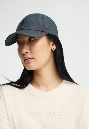 Young woman with long black hair wearing gold hoop earrings, a dark gray baseball cap, and a light beige shirt with embossed text.