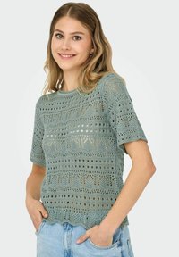 Suéter de crochet verde claro con mangas cortas, con patrones abiertos, un dobladillo en forma de concha y un acabado texturizado. Combinado con pantalones cortos de mezclilla.