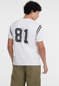 Hvid bomulds t-shirt med stort sort nummer "81" på ryggen, med sorte striber på skuldrene, iført med olivenfarvede cargo bukser.