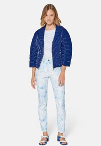 Marineblaue gesteppte Daunenjacke mit gekürzten Ärmeln über einem weißen Grafik-T-Shirt, kombiniert mit hellblauen Jeans im Marmor-Muster und blauen Sandalen.