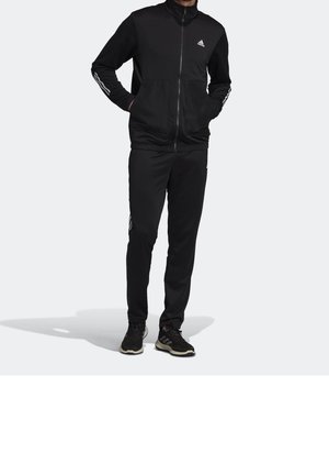 Personne portant un survêtement Adidas noir avec une veste zippée intégrale, des détails rayés sur les manches et le pantalon, et des baskets noires, debout les mains dans les poches.