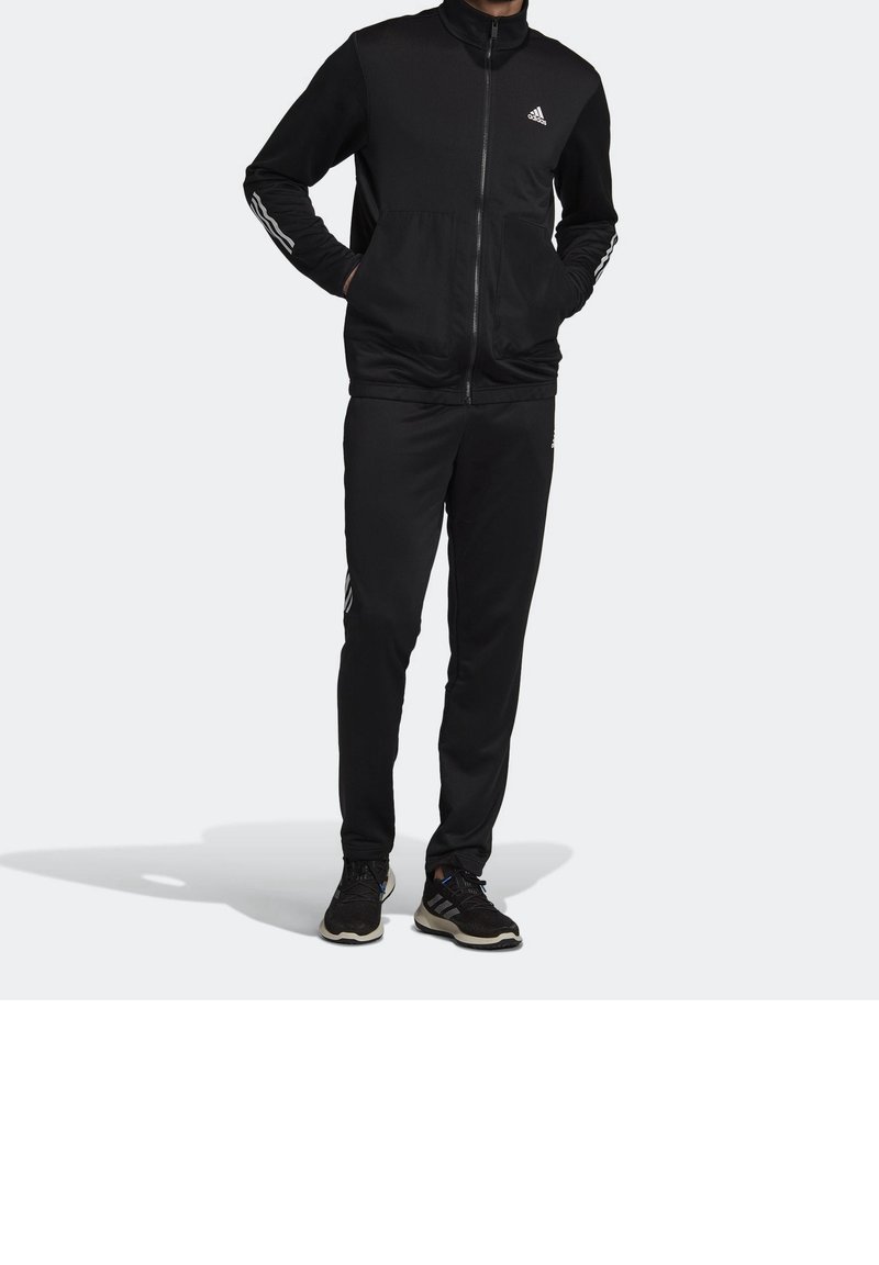 Personne portant un survêtement Adidas noir avec une veste zippée intégrale, des détails rayés sur les manches et le pantalon, et des baskets noires, debout les mains dans les poches.