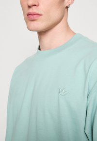 Sweatshirt verde mentă deschisă realizat dintr-un material moale, având un decolteu rotund și un mic logo brodat pe partea stângă a pieptului.