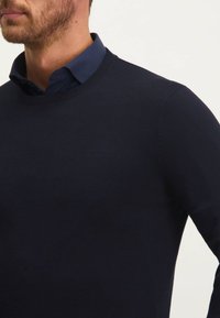 Marineblauer gestrickter Pullover mit Rundhalsausschnitt, der ein dezentes, geprägtes Logo auf der Brust und Ribbenbündchen aufweist. Glatte Textur und enganliegende Passform.