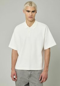 Polo shirt blanc avec col classique, manches courtes et coupe décontractée. Fabriqué en tissu doux, il présente une patte de boutonnage à trois boutons et un design simple.