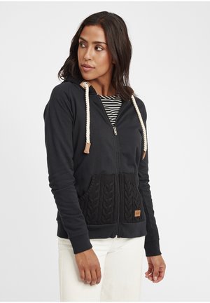 Felpa nera con zip caratterizzata da un design a tasca lavorata a maglia, cordini color crema e orlo a costine, indossata sopra una maglietta a righe con accenti beige.