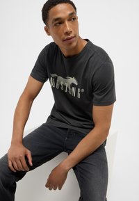 Camiseta de algodón gris oscuro con un logo de caballo y el texto "MUSTANG" en gris claro. Mangas cortas y diseño de cuello redondo.