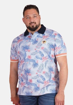 Homme aux cheveux courts et foncés avec une barbe, portant un polo à manches courtes à motifs floraux bleus et roses.