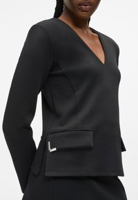 Egy nő fekete, testhez simuló, hosszú ujjú, V-nyakú felsőt visel, elöl zsebekkel, fehér háttér előtt.