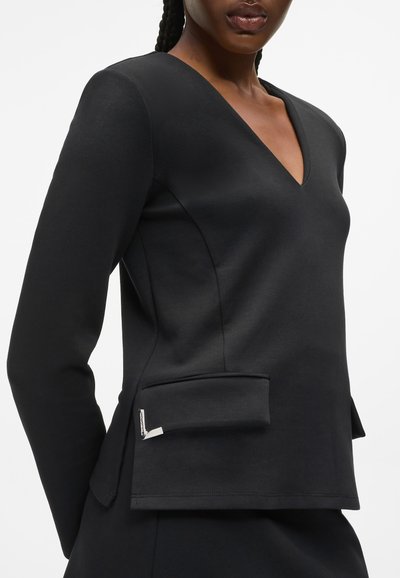 Femme portant un haut noir ajusté à manches longues avec un col en V et des poches à rabat à l'avant sur fond blanc.