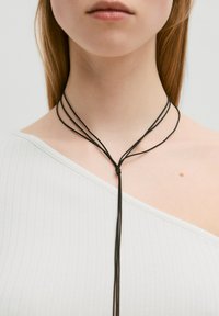 Zwarte koordketting met meerdere strengen, voorzien van een centrale knoop. Draag deze over een witte geribde top met een asymmetrische halslijn.
