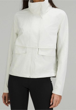 Vrouw draagt een wit jack met hoge kraag en klepzakken aan de voorkant, met een zwarte broek, staand tegen een effen grijze achtergrond.