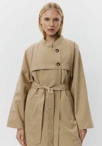 Beige trenčkot s vysokým límcem, zapínáním na dvojité knoflíky, páskem v oblasti pasu a širokými rukávy. Hladká textura, obsahující boční kapsy.