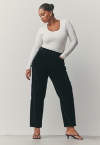 Hvid bodystocking med lange ærmer kombineret med højtaljede sorte fløjlsbukser og sorte strop-sandaler; glat tekstur og struktureret silhuet.