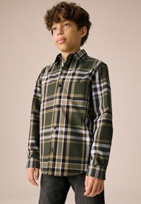Chemise à carreaux verts à boutons, avec des accents noirs et beige, dotée d'un col et de manches longues. Associée à un jean foncé.