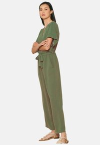 Mono verde oliva con mangas cortas y volantes, cintura ajustada, dos bolsillos laterales y corte holgado, combinado con sandalias ligeras de tiras.