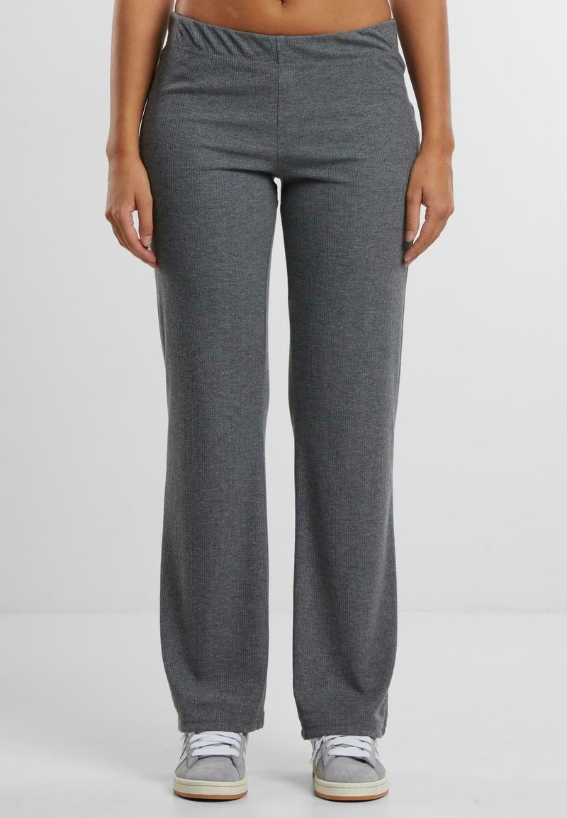 Personne portant un pantalon en maille gris taille haute à jambes droites avec des baskets grises et blanches à lacets, sur un fond uni.
