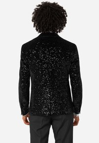 Oseba v črnem bleščečem sako in črnih hlačah, ki stoji s hrbtom obrnjenim proti gledalcu, na enobarvni beli podlagi.