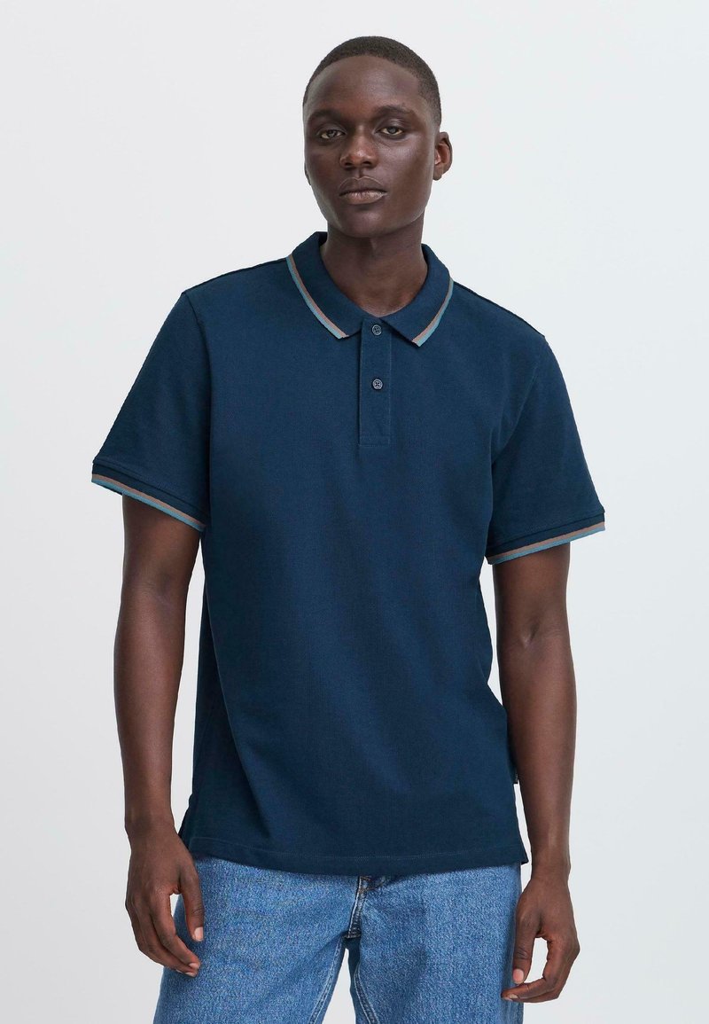 Mlad moški, oblečen v mornarsko modro kratkosleečno polo srajco z črtastim ovratnikom in robovi rokavov, v kombinaciji z svetlomodrimi jeans hlačami, stoji pred belo ozadje.