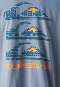 Lichtblauw T-shirt met een grafisch ontwerp van drie gestileerde golven en bergen in teal en oranje, met het woord "Quiksilver" in vet oranje.